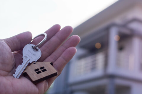 Eine Hand hält einen Schlüssel - im Hintergrund unscharf ein Haus.